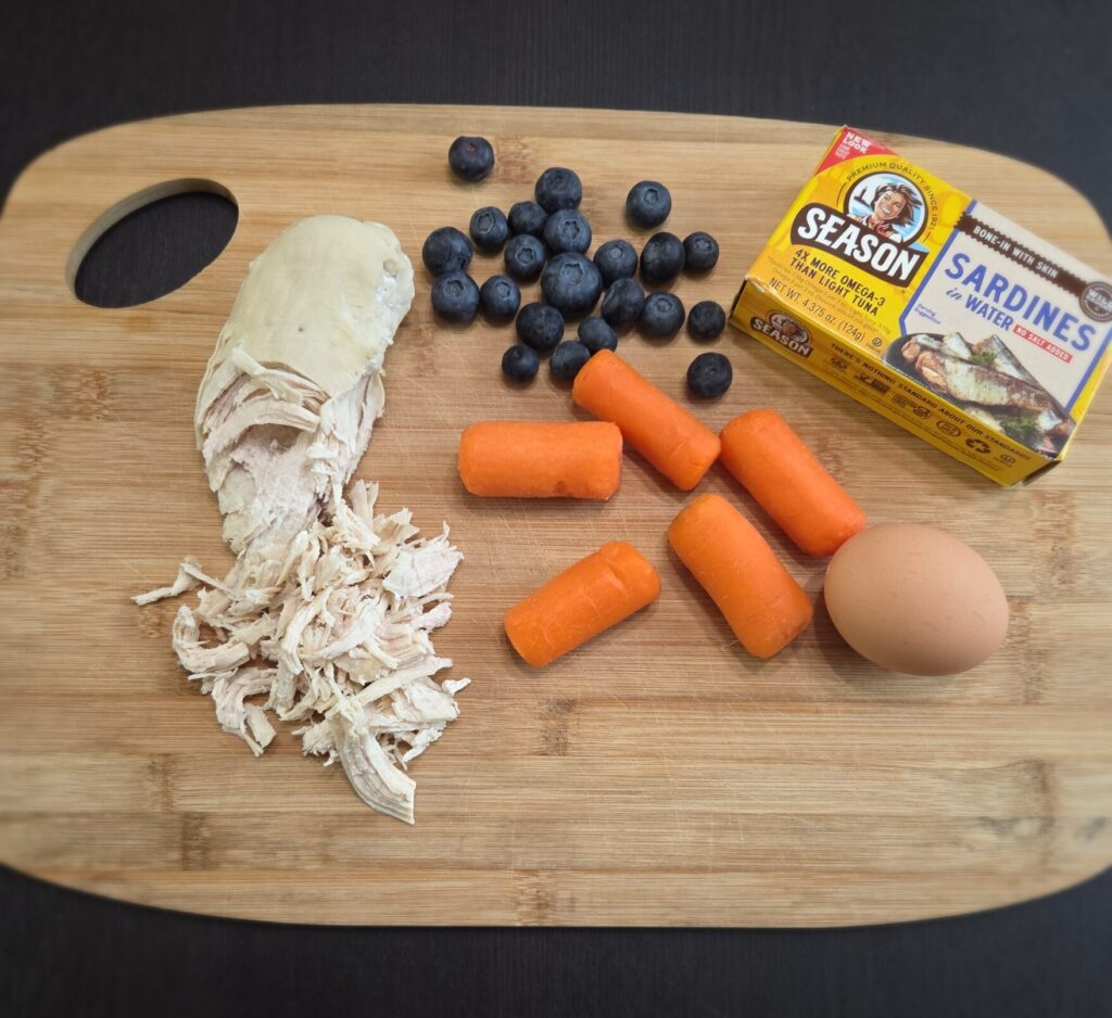 Fresh whole food ingredients for dogs including shredded chicken, blueberries, carrots, sardines, and egg on a wooden cutting board