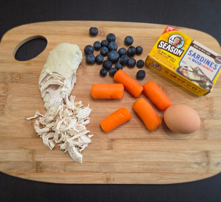 Fresh whole food ingredients for dogs including shredded chicken, blueberries, carrots, sardines, and egg on a wooden cutting board