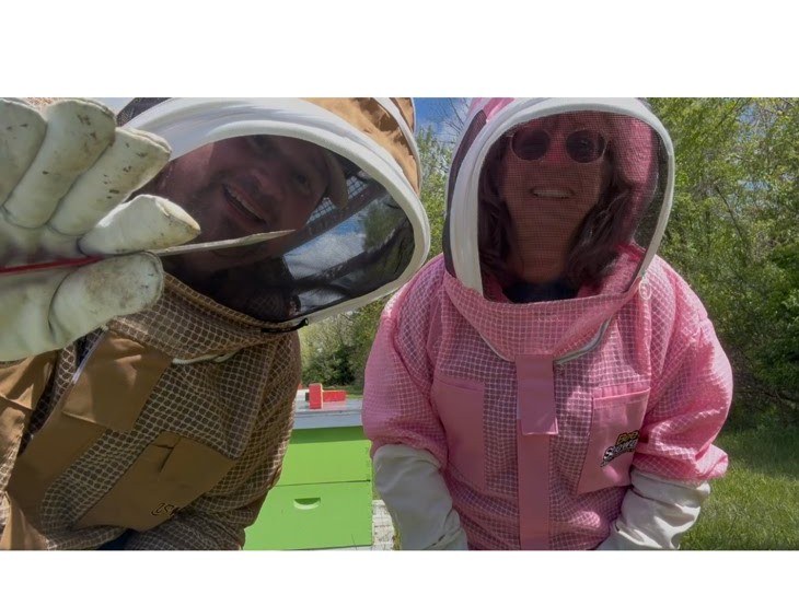 Connie Klar and beekeeper John Lytell in beekeeper suits waving at the camera at Bee Lyvely Farms in Indiana