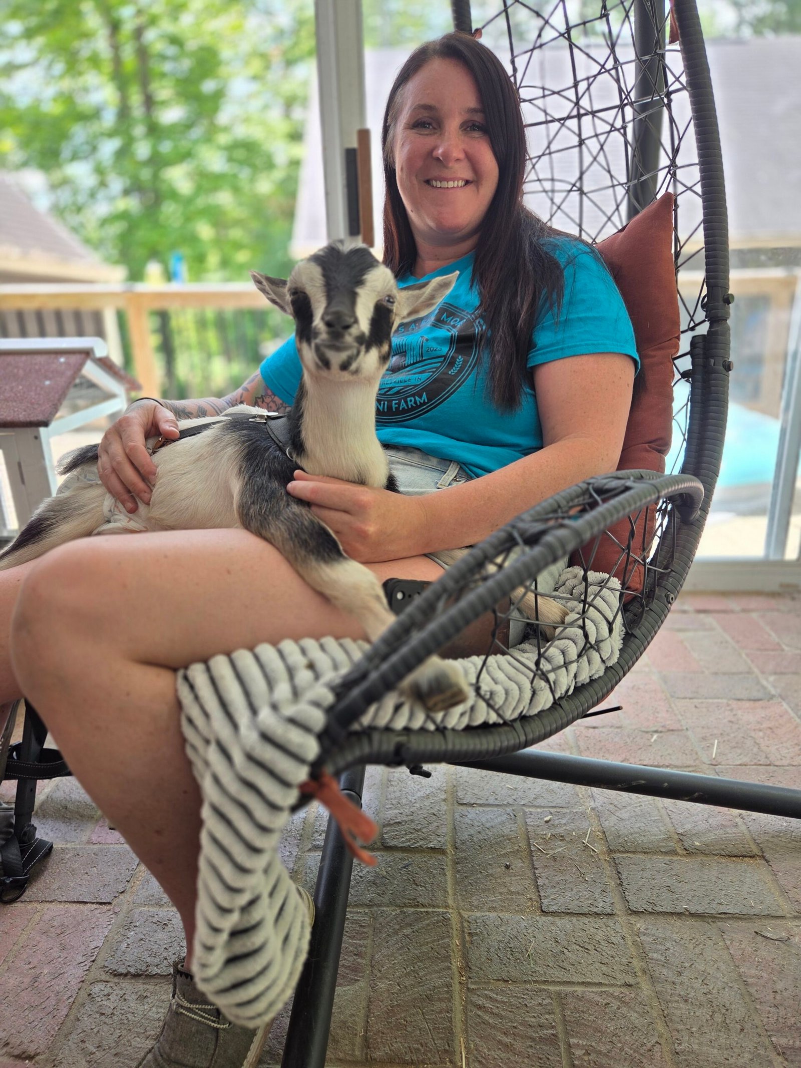 Cassie, founder of What's a Few More Mini Farm, holds Pippy the paralyzed Nigerian Dwarf goat in her lap while sitting in a hanging chair on her screened porch in Mooresville, Indiana.